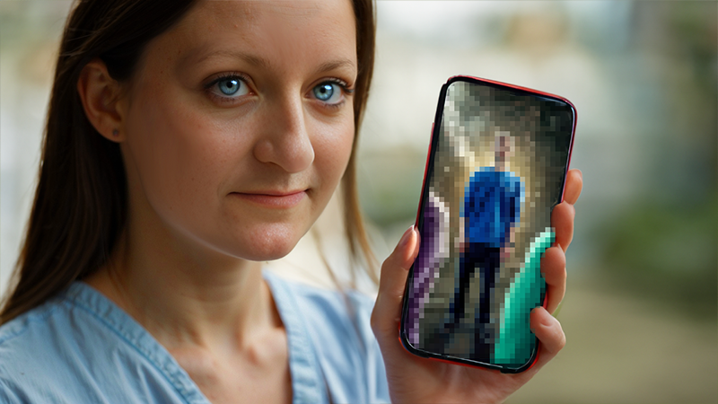 Femme (deepfake de Flore du Web, donc moi-même), tenant un smartphone à écran pixellisé, regardant l'objectif avec un sourire discret.